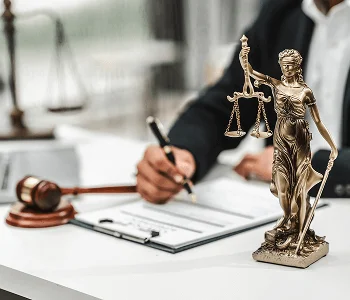 A golden Lady Justice statue stands beside a gavel and a person signing legal documents at a desk.